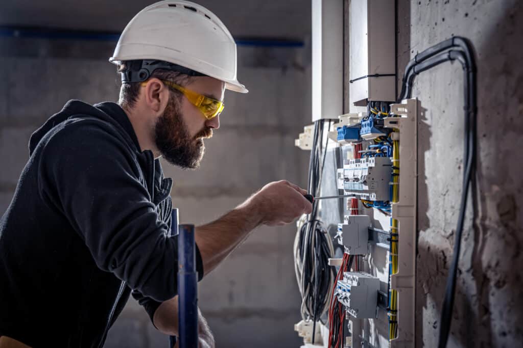 Électricien barbu avec casque blanc et lunettes de sécurité jaunes vissant un disjoncteur dans un panneau électrique moderne sur chantier.