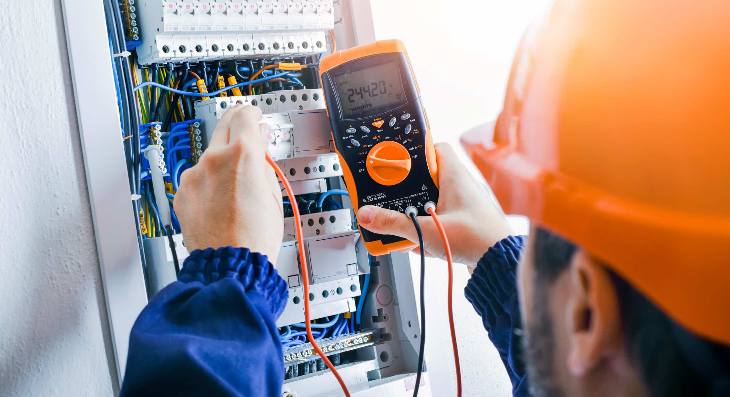 Électricien mesurant la tension (244.20 V) d'un tableau électrique avec un multimètre. Disjoncteurs B 16 visibles.
