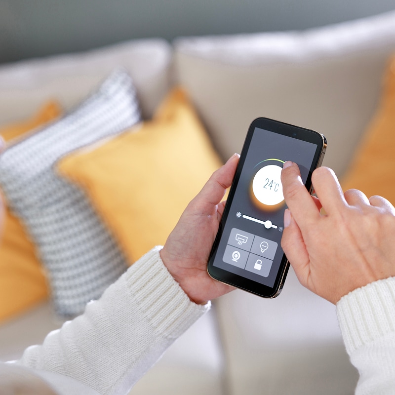 Mains réglant un thermostat intelligent à 24°C via une application mobile sur smartphone, avec des coussins jaunes en arrière-plan.