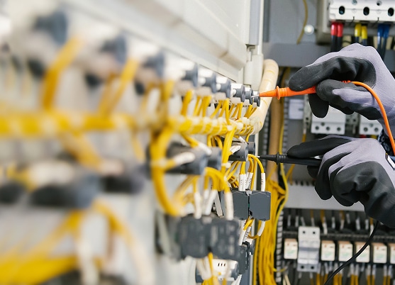 Électricien en gants gris testant des connexions électriques jaunes dans un tableau de commande avec un multimètre.