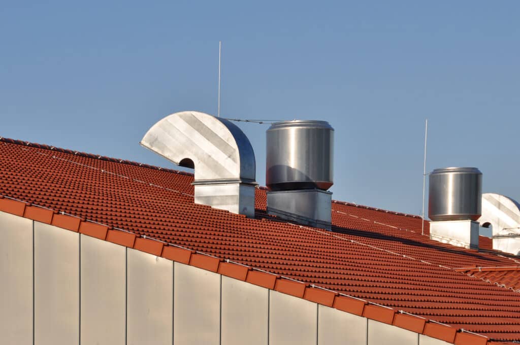 Toit de tuiles rouges avec de grands conduits de ventilation métalliques brillants et des paratonnerres sous un ciel bleu clair.