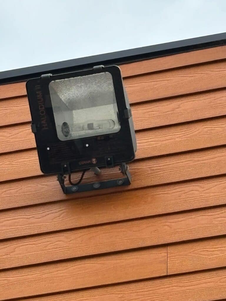 Projecteur Extérieur OSRAM Halodium II sur Mur en Bois Projecteur OSRAM Halodium II fixé sur un mur en bois brun, luminaire extérieur avec réflecteur et ampoule visibles.